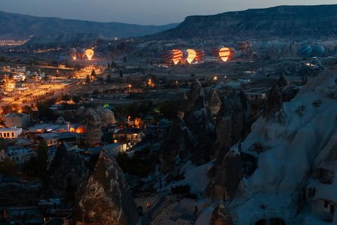 Göreme in Kappadokien zur blauen Stunde, beleuchtete Häuser und Felskegel, dahinter aufsteigende Heißluftballons