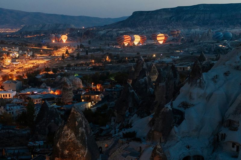 Göreme in Kappadokien zur blauen Stunde, beleuchtete Häuser und Felskegel, dahinter aufsteigende Heißluftballons