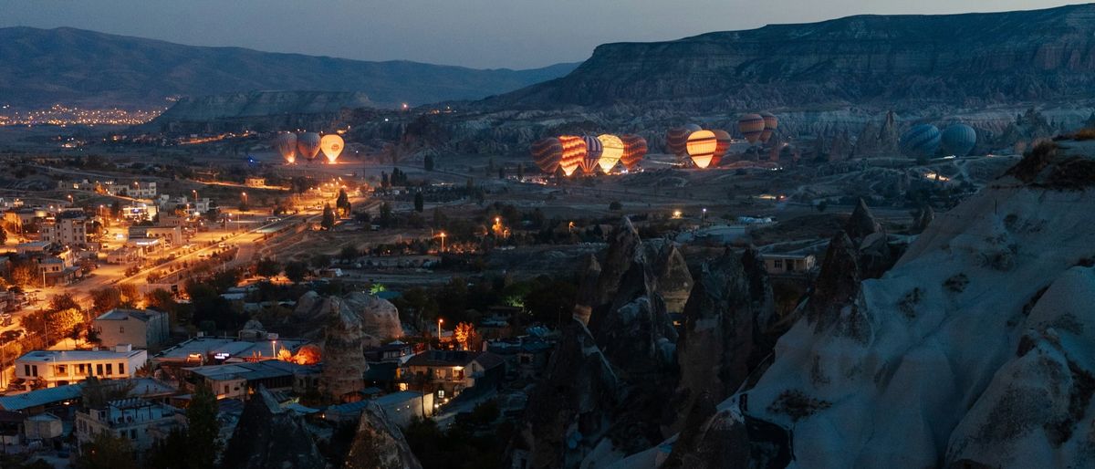 Göreme in Kappadokien zur blauen Stunde, beleuchtete Häuser und Felskegel, dahinter aufsteigende Heißluftballons