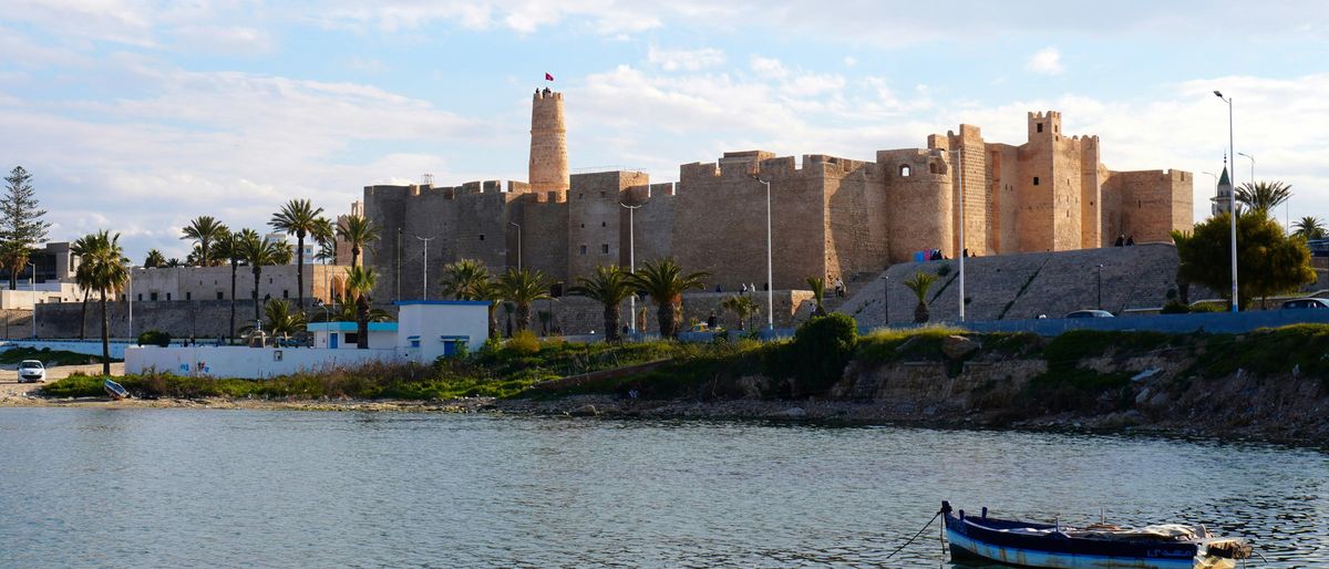 Ribat von Monastir über einer ruhigen Bucht, davor Palmen und ein kleines blaues Boot auf dem Wasser
