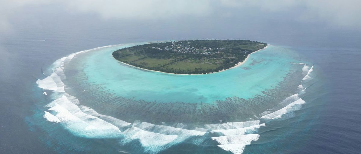 Die bewohnte Insel Thoddoo auf den Malediven aus der Luft: Dorf, Felder, türkise Lagune und Brandung am Riff unter Wolken